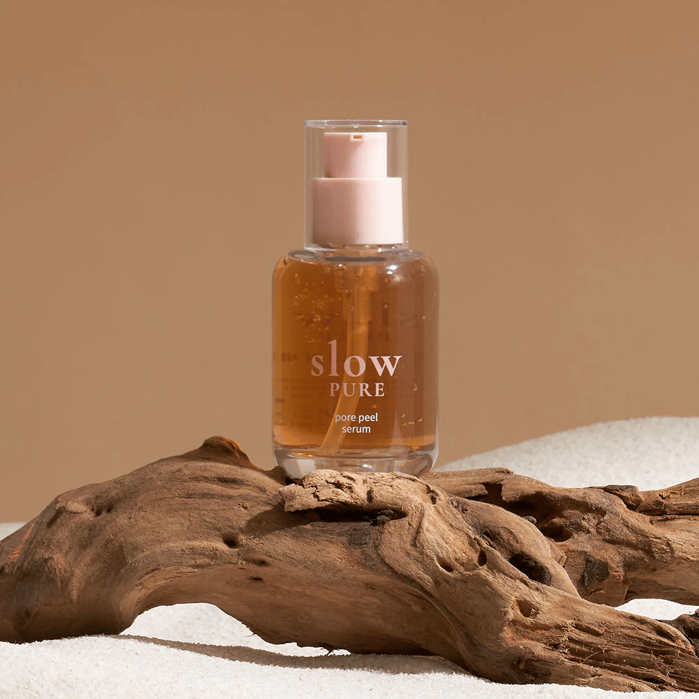 Slow Pure Pore Peel Serum geplaatst op zand met houten detail op de achtergrond, symbool voor natuurlijke huidverzorging.