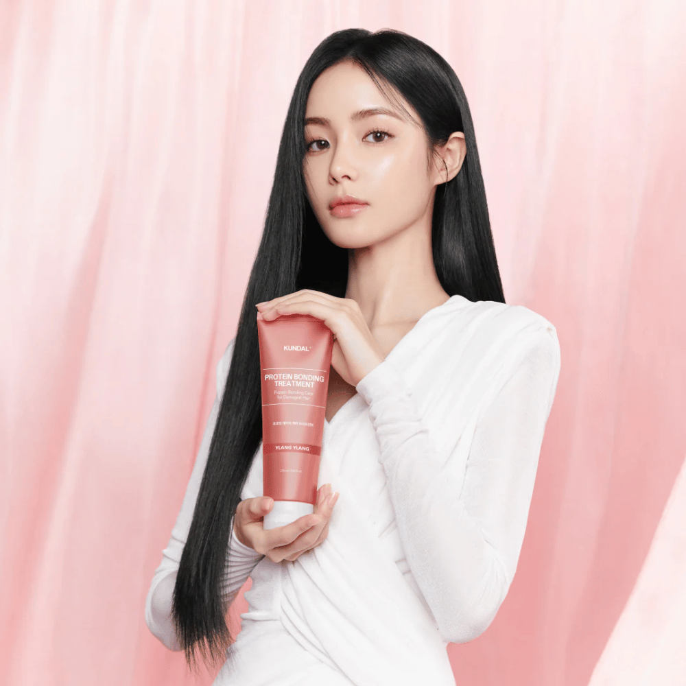 Vrouw met lang, glanzend haar houdt KUNDAL Protein Bonding Treatment Violet Muguet tegen roze achtergrond.