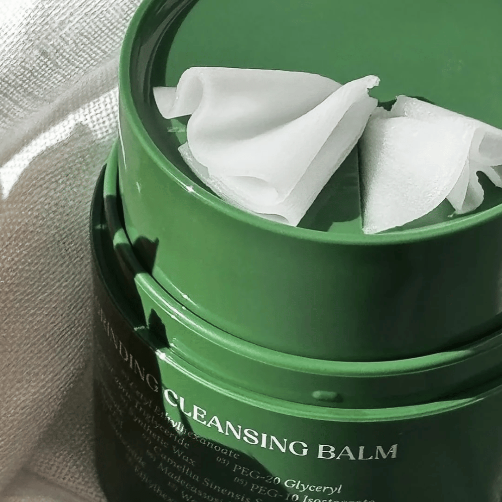 De afbeelding toont een groene pot met "Cleansing Balm", die geopend is om de bijgeleverde spatel en de zachte inhoud te onthullen. De romige textuur weerkaatst het licht, wat een gevoel van luxe en efficiëntie in de reiniging geeft.