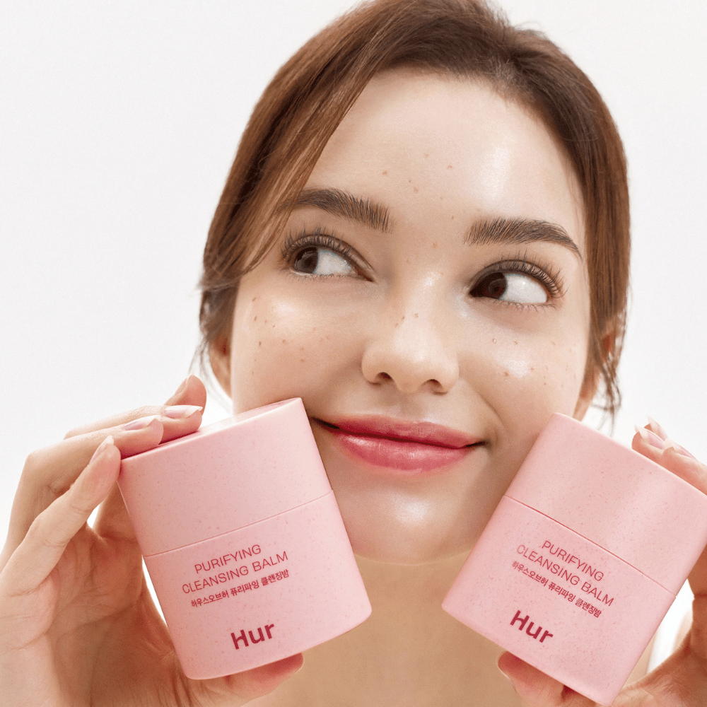 Model houdt twee potjes Hoe Purifying Cleansing Balm. Milde reinigingsbalsem die geschikt is voor gevoelige huid en een frisse, gehydrateerde uitstraling na reiniging geeft.