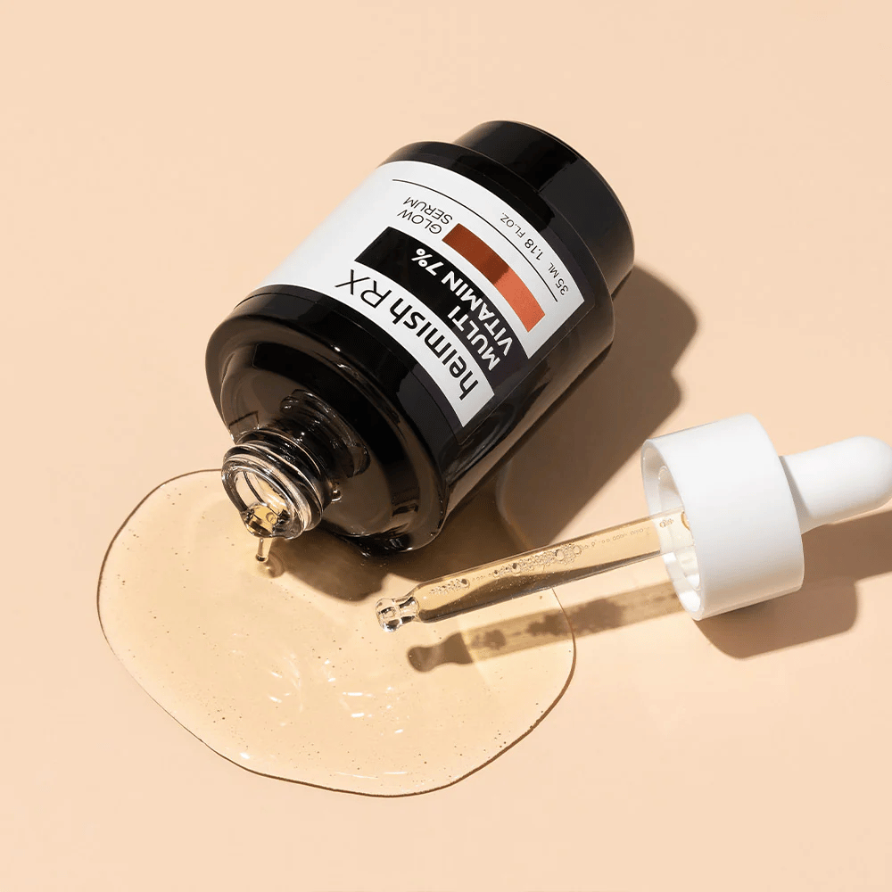 Heimish RX Multi Vitamin 7% Glow Serum liggend op een beige achtergrond met serum verspreid rond de fles en een pipet gevuld met product, wat de voedzame formule benadrukt.