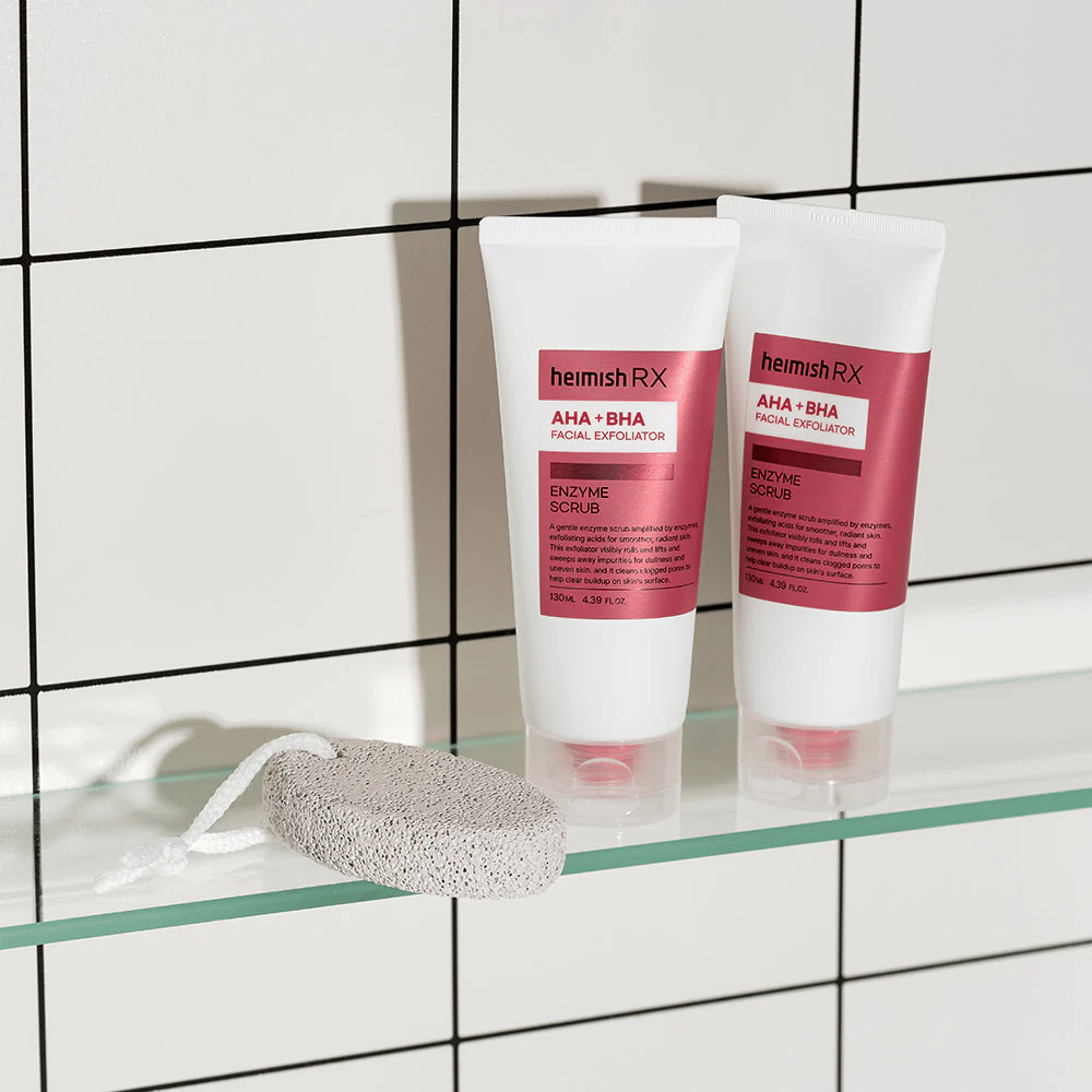 Twee tubes Heimish RX AHA + BHA Facial Exfoliator geplaatst op een glazen plank in een badkamer met witte tegelmuur. Naast de producten ligt een puimsteen met een wit touwtje. Een stijlvolle en minimalistische huidverzorgingsfoto.