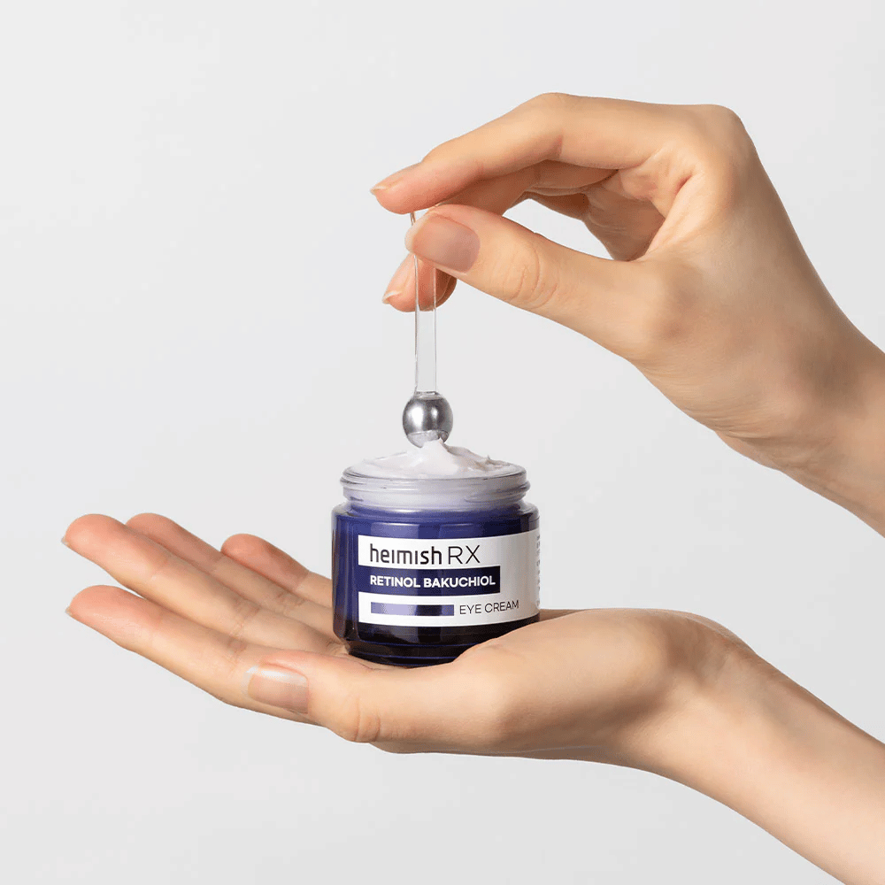 Heimish RX Retinol Bakuchiol Eye Cream in een elegante blauwe pot, opgetild met een metalen applicator. Een oogcrème met retinol en bakuchiol om fijne lijntjes te verminderen en een stevigere huid te geven.