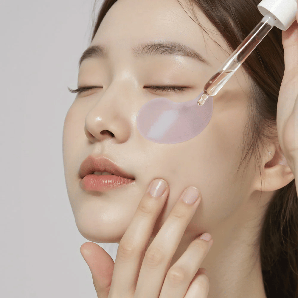 Een vrouw met een paarse hydrogel-ooglap brengt serum aan onder het oog met een pipet. Het product hydrateert en verzorgt de gevoelige huid rond de ogen.