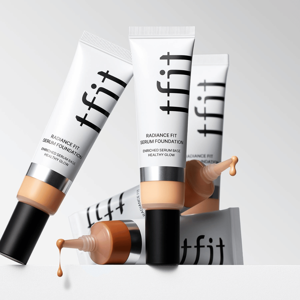 Meerdere tubes van TFIT Radiance Fit Serum Foundation in verschillende tinten, waarvan één met foundation die eruit druppelt, tegen een lichtgrijze achtergrond.