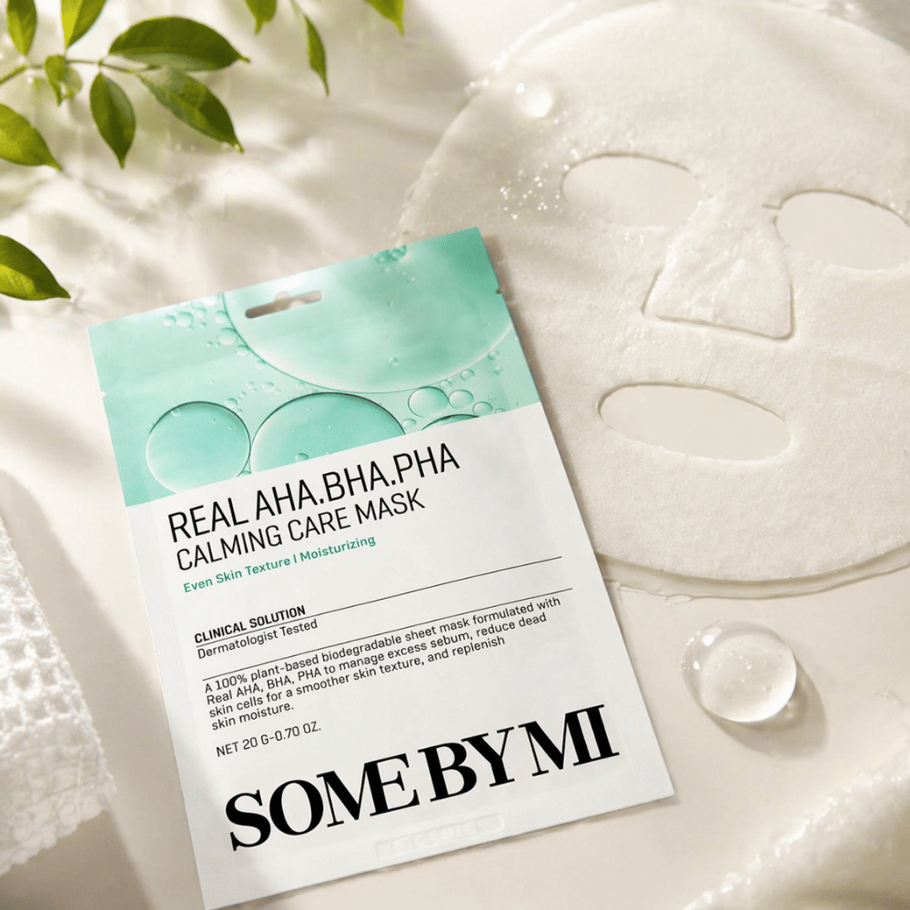 SOME BY MI Real AHA.BHA.PHA Calming Care Mask wordt getoond samen met het ontvouwde sheetmasker. Rond het product zijn vochtdruppels en groene bladeren te zien die het gevoel van hydratatie, kalmerende verzorging en milde exfoliatie versterken. De afbeelding benadrukt de focus van het masker op een egalere huidtextuur en een verbeterde vochtbalans.