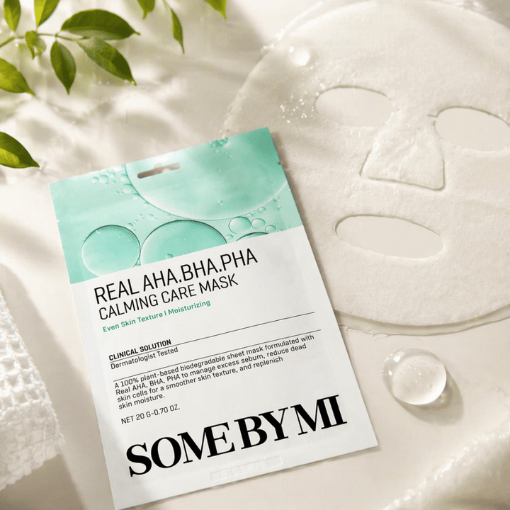 SOME BY MI Real AHA.BHA.PHA Calming Care Mask wordt getoond samen met het ontvouwde sheetmasker. Rond het product zijn vochtdruppels en groene bladeren te zien die het gevoel van hydratatie, kalmerende verzorging en milde exfoliatie versterken. De afbeelding benadrukt de focus van het masker op een egalere huidtextuur en een verbeterde vochtbalans.