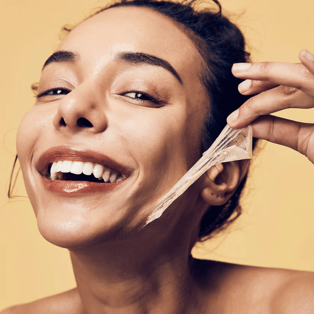De afbeelding toont een vrouw met een grote glimlach die bezig is een peel-off masker van haar gezicht te verwijderen. De vrouw ziet er blij en tevreden uit terwijl ze het schoonheidsproduct gebruikt, wat een gevoel van welzijn en huidverzorging overbrengt.