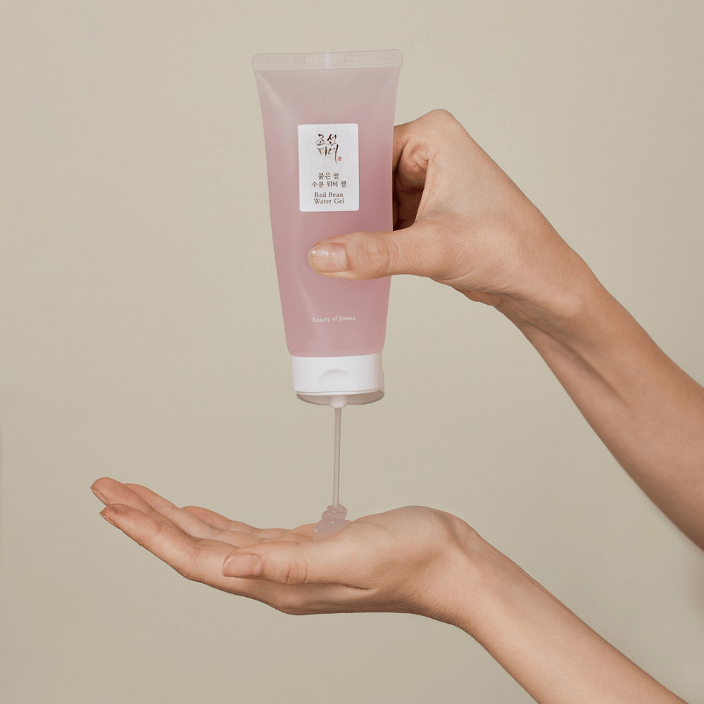 De afbeelding toont een persoon die een tube van "Red Bean Water Gel" van Beauty of Joseon vasthoudt en een doorzichtige gel in zijn hand knijpt. Het product lijkt bedoeld te zijn voor huidverzorging en heeft een aantrekkelijke, zachtroze verpakking.
