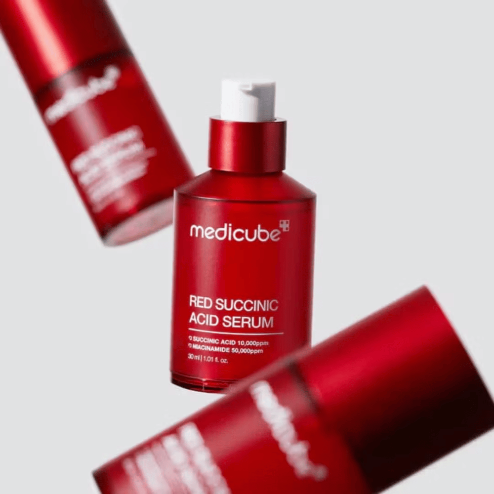 Medicube Red Succinic Acid Serum in een rode pompflacon, omringd door meer identieke flessen tegen een lichte achtergrond. Het product bevat succinic acid en niacinamide om acne en pigmentvlekken te behandelen.