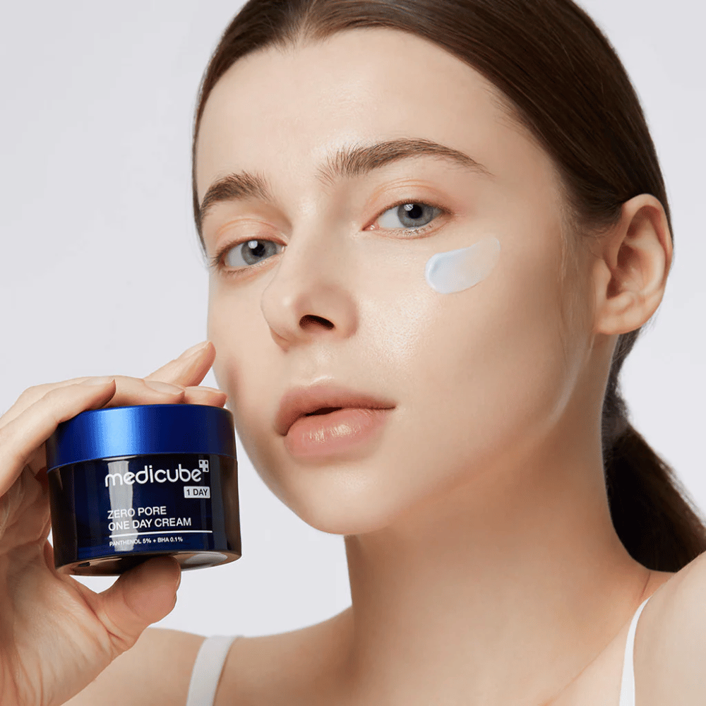 Vrouw met een natuurlijke look houdt Medicube Zero Pore One Day Cream. Een kleine hoeveelheid crème is op haar wang aangebracht. Het product is ontworpen om de huidstructuur te verbeteren en de poriegrootte te minimaliseren, met een lichte en hydraterende formule.