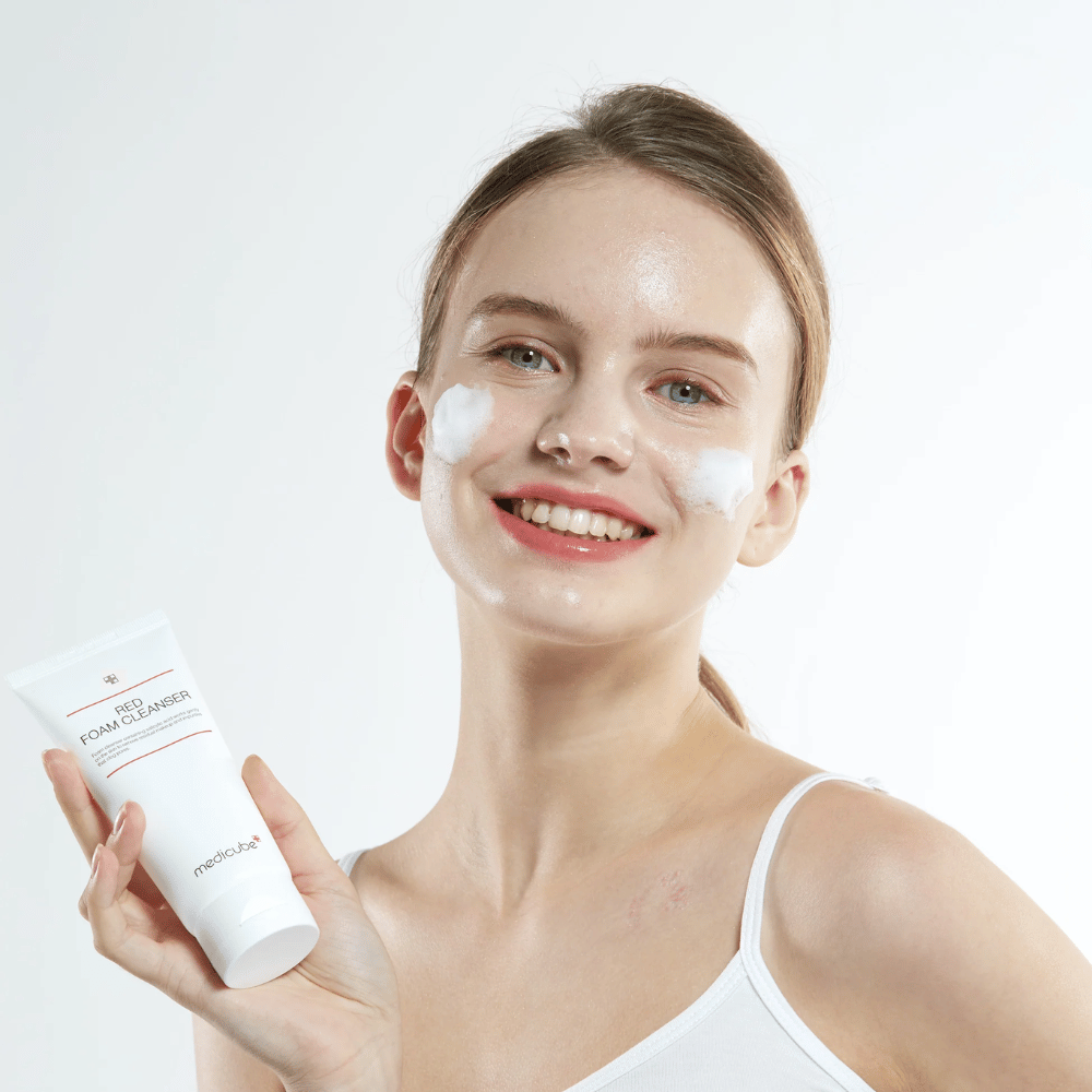 Ung kvinna med rengöringsskum i ansiktet håller upp en tub av Medicube Red Foam Cleanser och ler mot kameran. Hon bär ett vitt linne och bakgrunden är ljus och enkel.