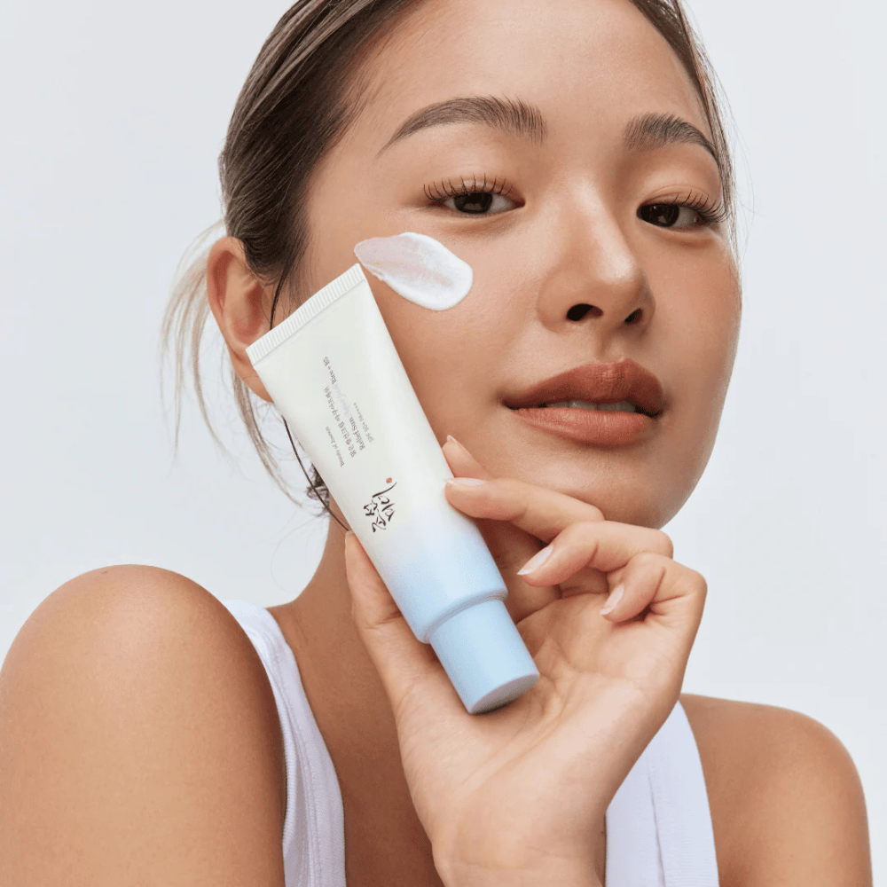 Vrouw houdt Beauty of Joseon Relief Sun: Aqua-Fresh Rice + B5 zonnebrandcrème en toont de romige textuur op de wang. Een lichte zonnebeschermingsproduct die hydrateert en de huid beschermt met SPF 50+ PA++++.