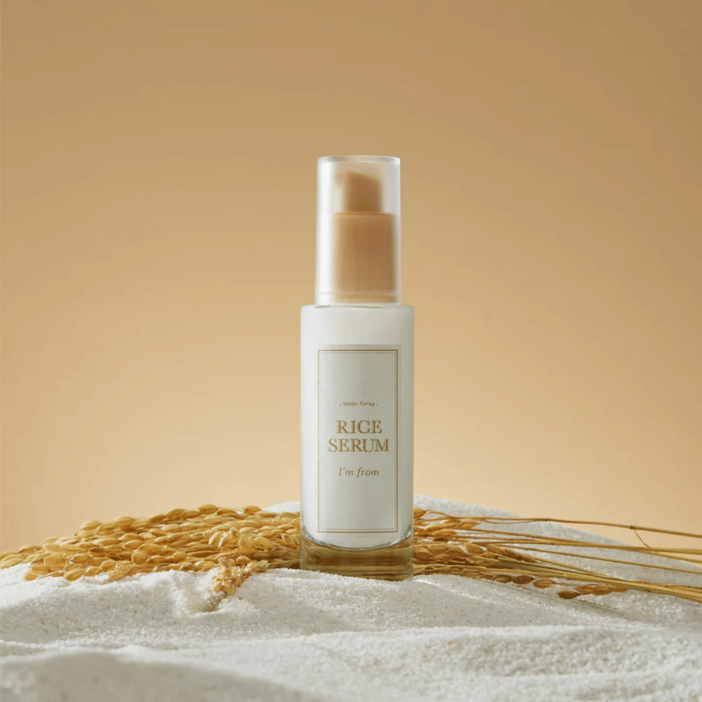 Fles met "I'm from" Rice Serum, omgeven door rijstkorrels en rijsthalmen tegen een warme achtergrond.