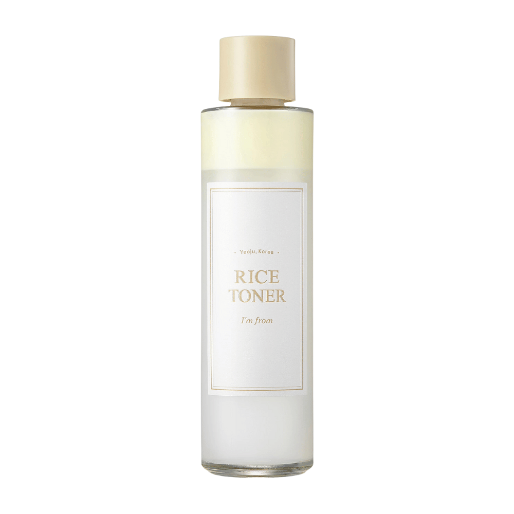 Een transparante fles met een gouden dop en een etiket dat aangeeft "Rice Toner I'm from Yeojü, Korea". De fles bevat een lichte vloeistof, waarschijnlijk toner op basis van rijstextract. Het ontwerp is schoon en minimalistisch. De achtergrond is wit.