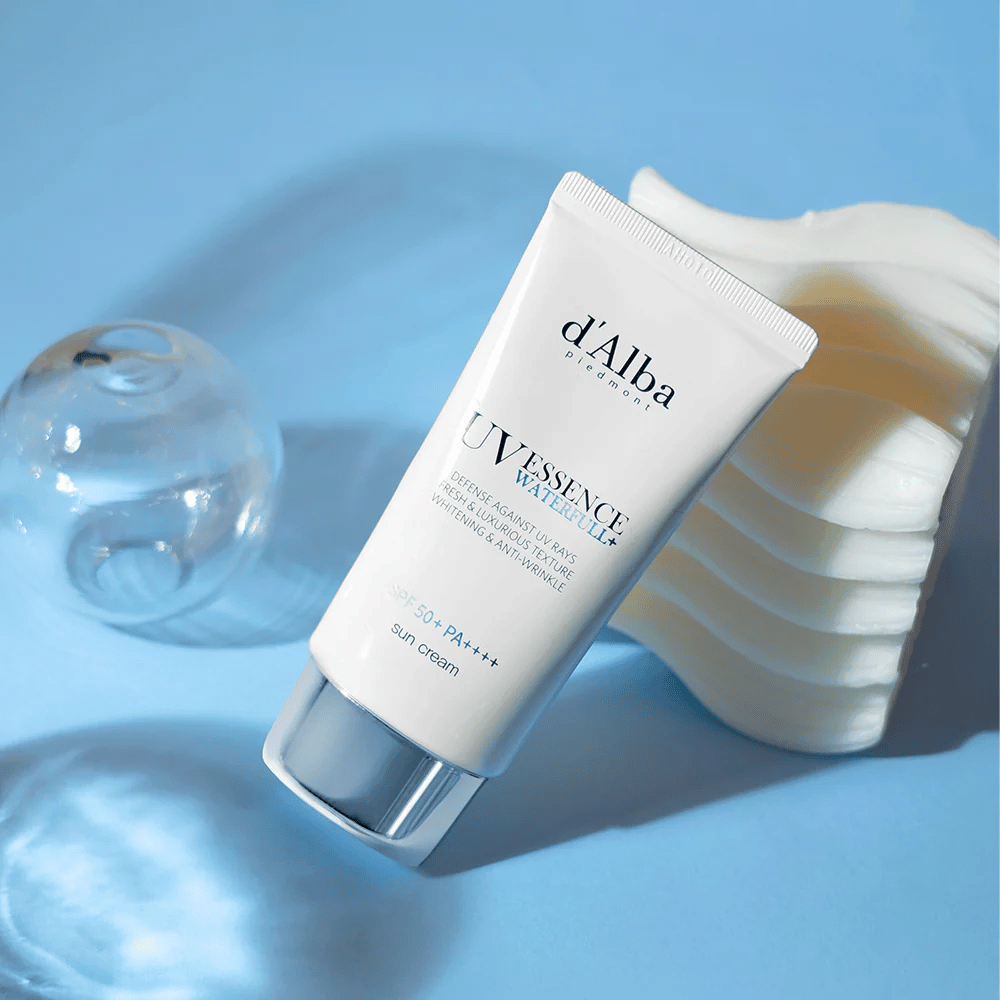 d'Alba UV Essence Waterfull+ Sun Cream met SPF 50+ PA++++ ligt tegen een blauwe achtergrond met lichtreflecties en decoratieve elementen - benadrukt de frisse en hydraterende eigenschappen van de zonnecrème.