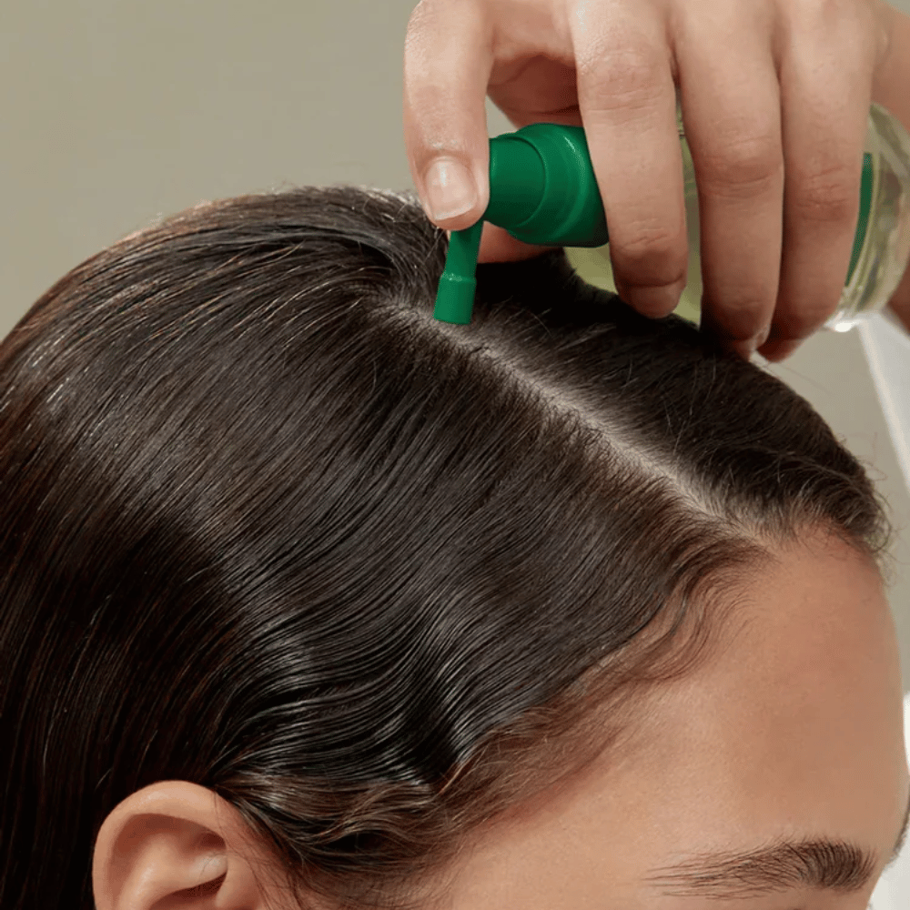Persoon brengt AROMATICA Rosemary Root Enhancer rechtstreeks aan op de hoofdhuid met behulp van het gerichte mondstuk. De afbeelding toont hoe het product wordt gebruikt voor hoofdhuidverzorging en versterking van de haarwortels.