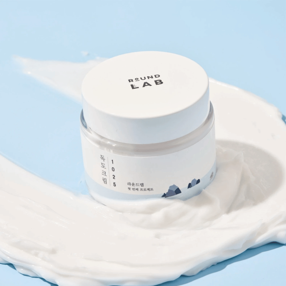 Een pot met Round Labs 1025 Dokdo Cream 80 ml die hydratatie biedt, geplaatst op een levendige blauwe achtergrond.