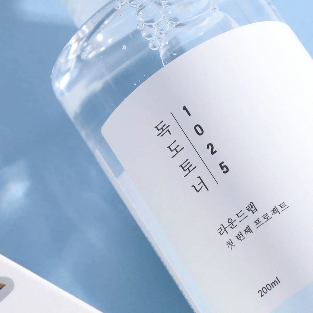 Close-up van een doorzichtige fles Round Lab 1025 Dokdo Toner, gevuld met heldere vloeistof, tegen een blauwe achtergrond.