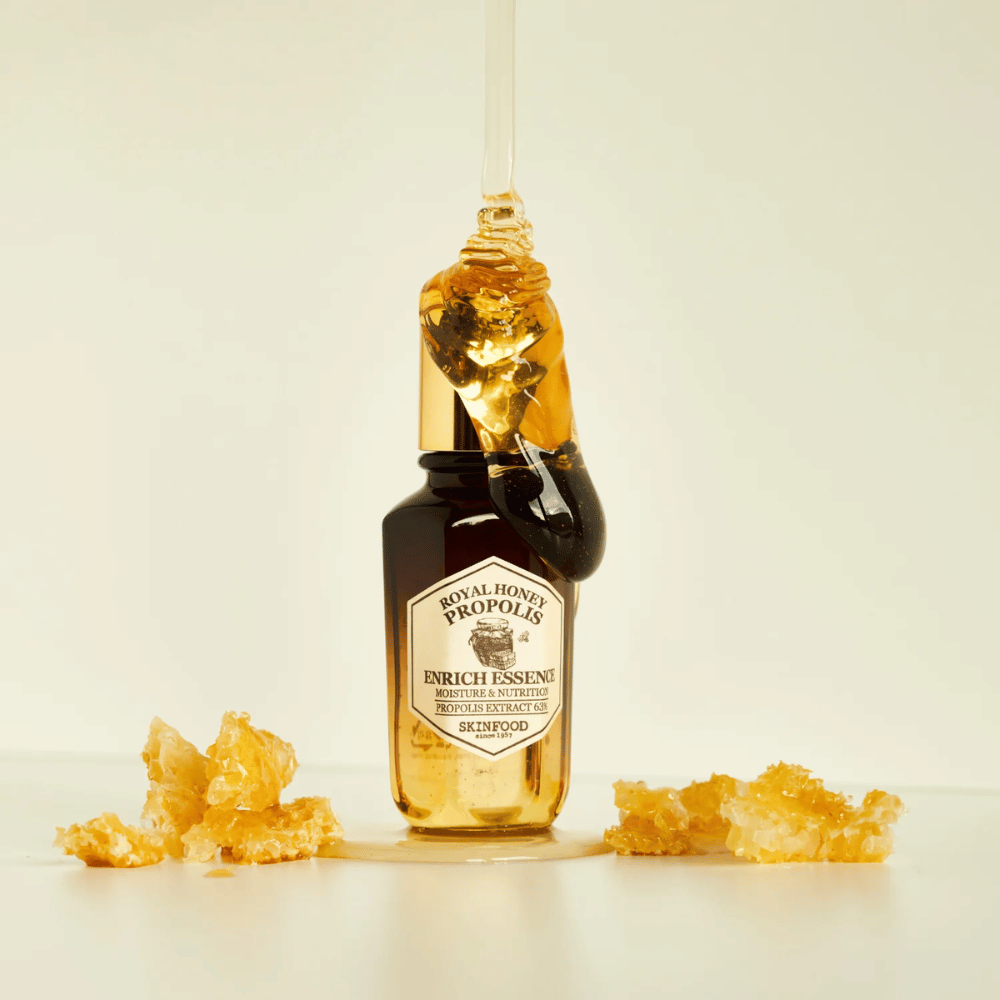 Afbeelding van een glazen fles met Royal Honey Propolis Enrich Essence van Skinfood. Een grote druppel gouden honing loopt van de goudkleurige dop van de fles. Rond de fles liggen kleine stukjes honingraat verspreid op een lichte ondergrond. De essentie wordt op de markt gebracht met de nadruk op vocht en voeding, met 63% propolisextract.