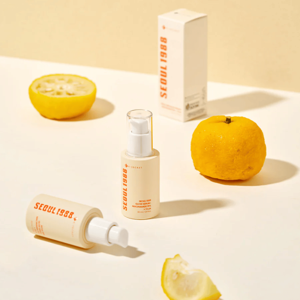 Meerdere flessen en verpakkingen van Seoul 1988 Glow Serum gerangschikt met hele en halve yuja-vruchten op de achtergrond.