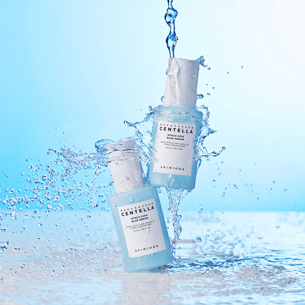 Twee flessen SKIN1004 Madagascar Centella Hyalu-Cica Blue Serum worden geraakt door een waterval tegen een lichtblauwe achtergrond.