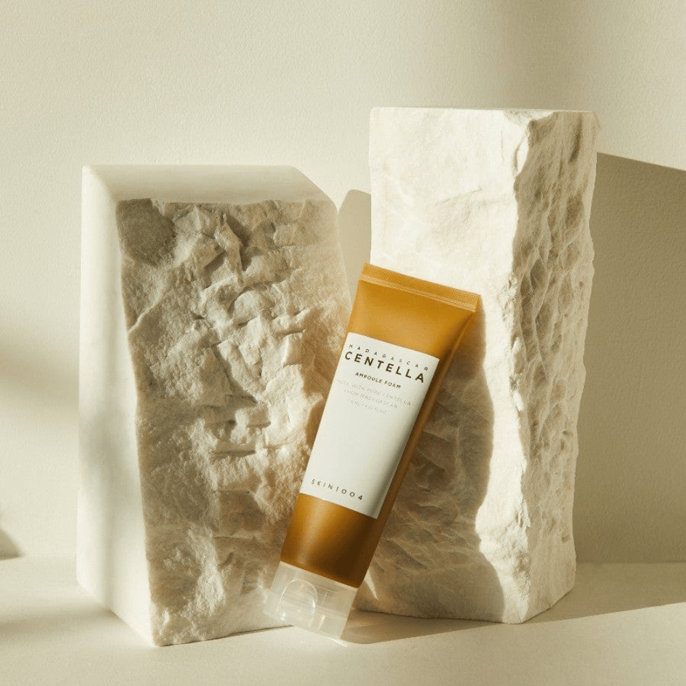 SKIN1004 Madagascar Centella Ampoule Foam staande tegen lichte steenblokken in natuurlijk zonlicht, met hun bruin-witte tubedesign en elegante minimalisme.