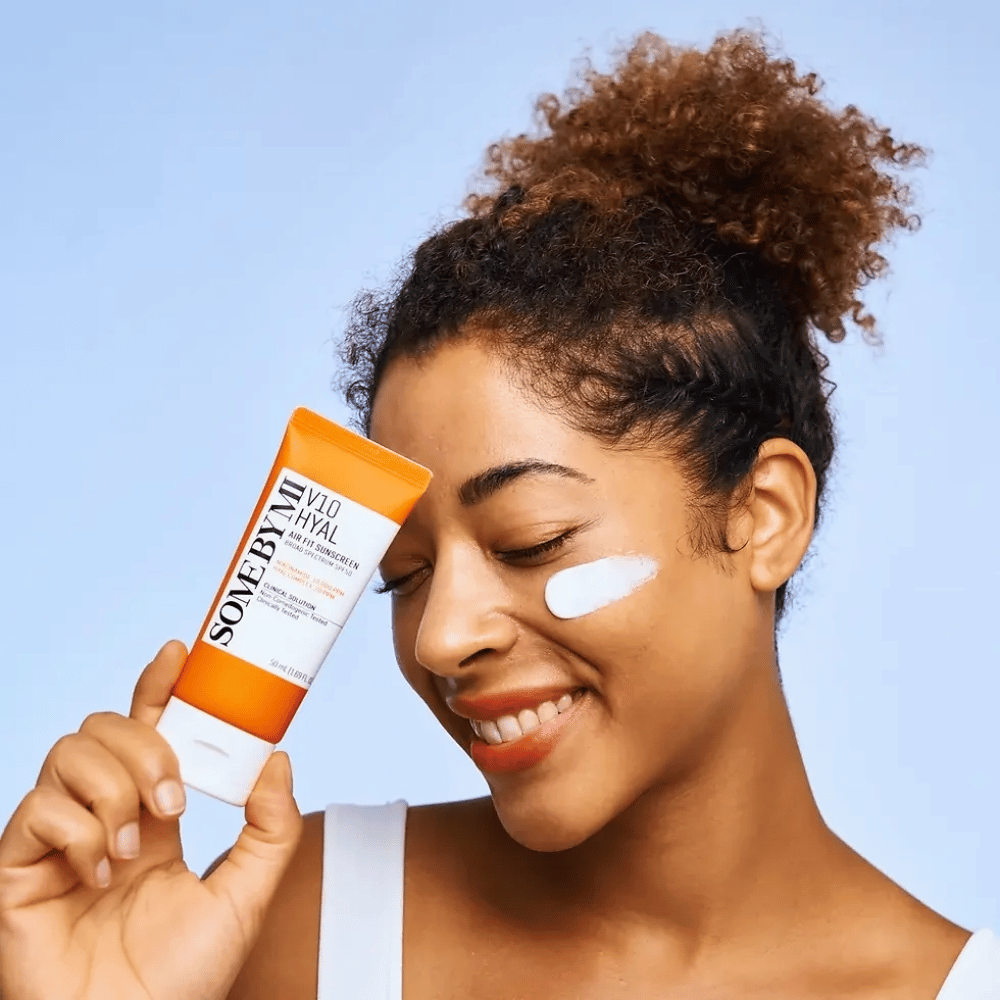 Een vrouw houdt een tube met SOME BY MI V10 Hyal Air Fit Sunscreen en heeft een witte vlek zonnebrandcrème op haar wang. Ze glimlacht vrolijk en toont duidelijk het product, wat de toepassing en voordelen voor huidverzorging en bescherming tegen de zon benadrukt.
