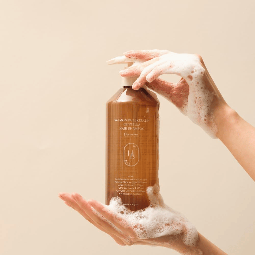 Heveblue Salmon Pullkeratin Centella Hair Shampoo in een bruine fles omringd door schuim. Het product wordt tussen twee handen vastgehouden en benadrukt een reinigende en verzorgende haarverzorgingservaring met rijkelijk schuim.