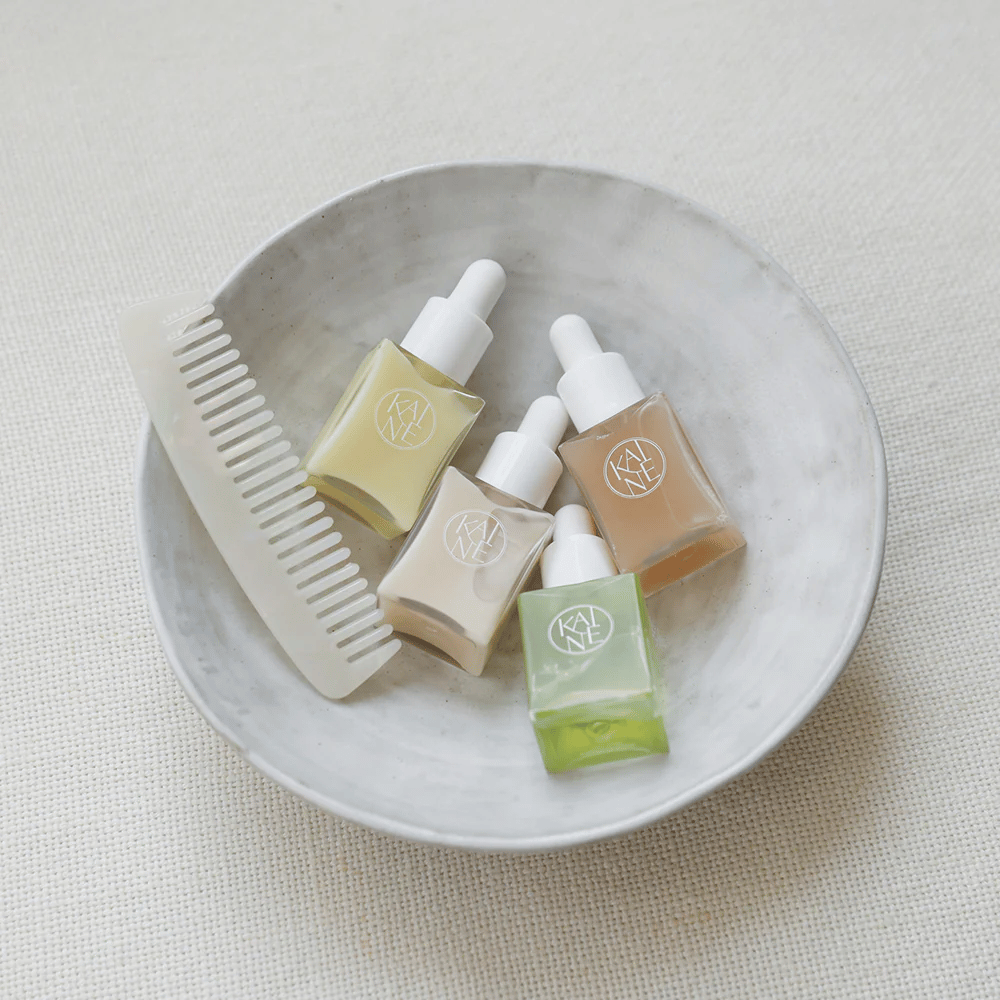 Vier kleine serumflessen van KAINÉ Serum Discovery Kit liggen in een witte schaal naast een kam, met een natuurlijke en rustige achtergrond in een beige tint.