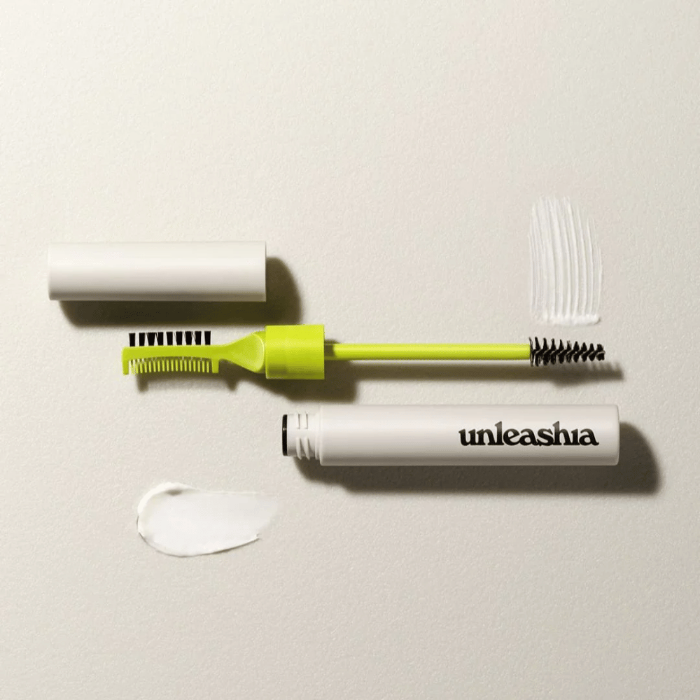 Een wenkbrauwsculptuurset met een groene kam, zwarte borstel en een witte tube gemarkeerd "unleashia" op een lichte achtergrond met een klomp van transparant product.