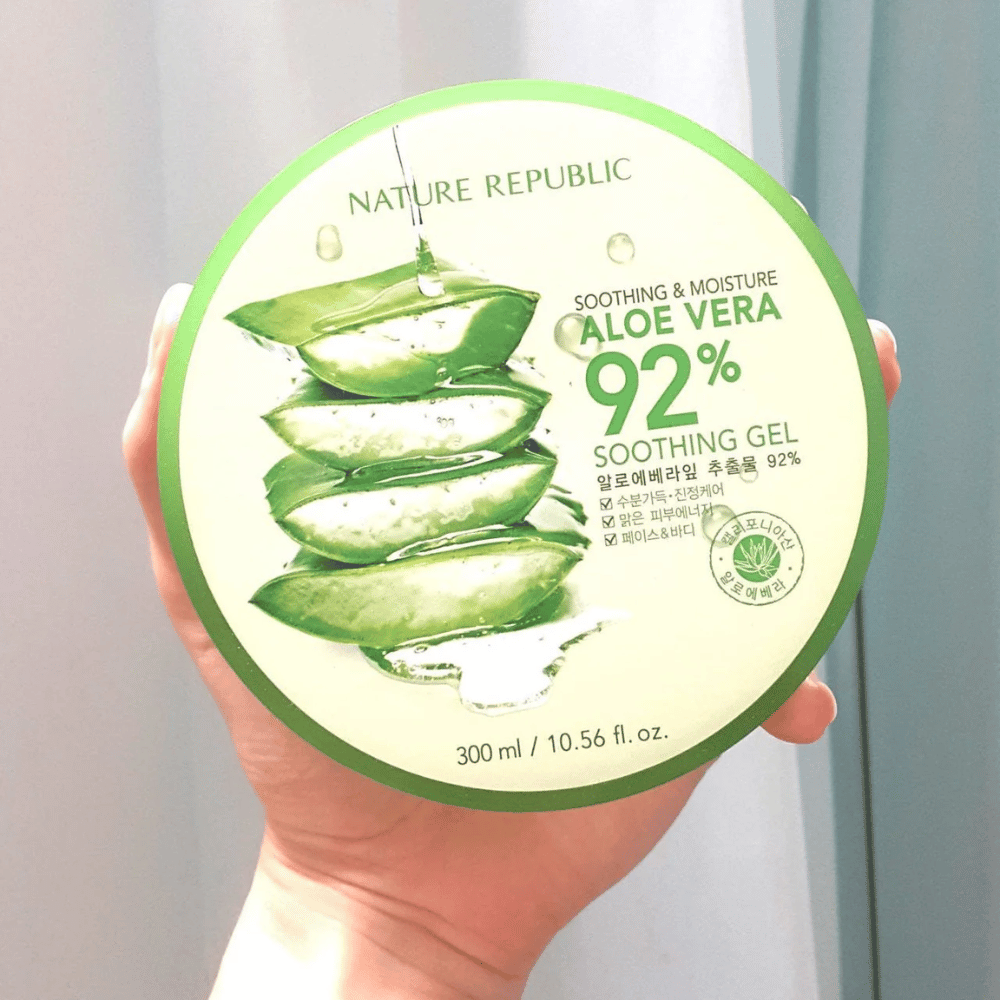 Een hand houdt een ronde pot met Nature Republic Aloe Vera 92% Verzachtende Gel omhoog. De pot is groen met een afbeelding van aloë vera en tekst die de verzachtende en hydraterende eigenschappen beschrijft. De inhoud is 300 ml.