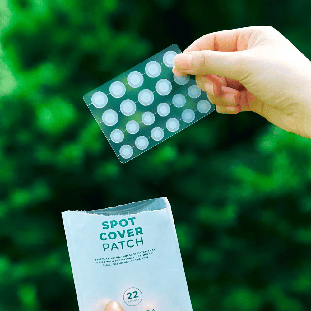 Een hand houdt een vel Spot Cover Patches omhoog, die transparant en afgerond zijn. Op de achtergrond is een wazige groene omgeving te zien. De verpakking toont een deel van de verpakking met de tekst "SPOT COVER PATCH" en informatie dat het product 22 pleisters bevat die helpen kleine huidgebieden te genezen.