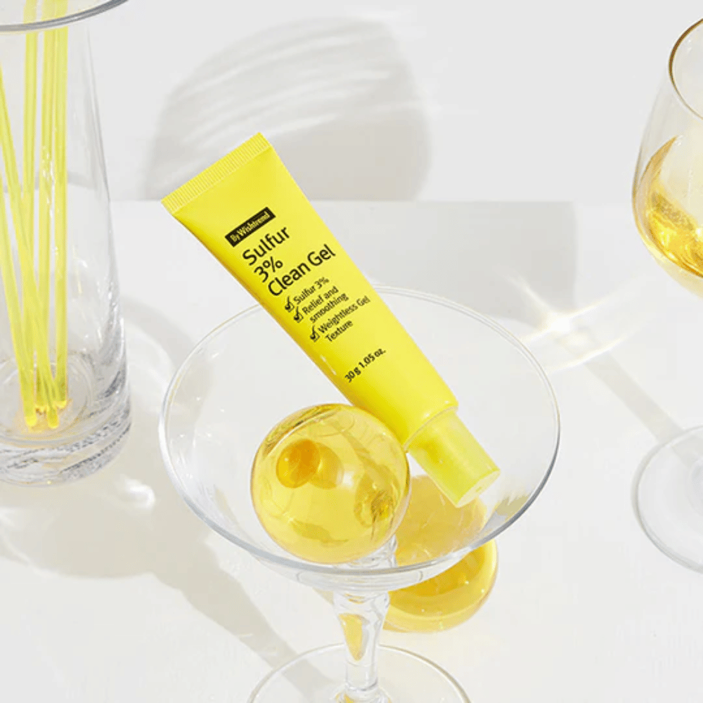 By Wishtrend Sulfur 3% Clean Gel in een gele tube geplaatst in een cocktailglas met een gele bol, omringd door elegante glazen op een witte achtergrond.
