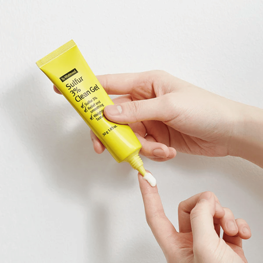 Close-up van handen die By Wishtrend Sulfur 3% Clean Gel aanbrengen, een gele tube met witte gel, tegen een witte achtergrond.