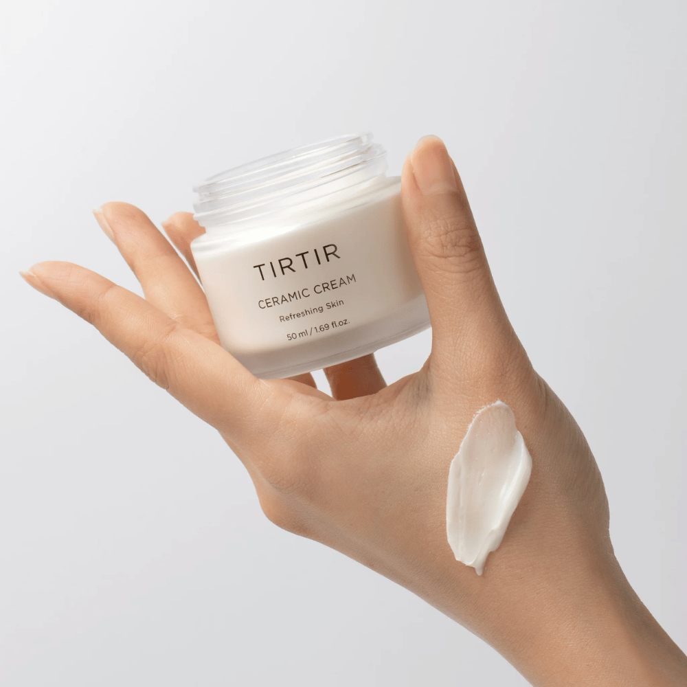 Een hand houdt een pot met TIRTIR Ceramic Cream vast. Op de rug van de hand is een hoeveelheid van de crème aangebracht. De crème lijkt dik en hydraterend te zijn. De verpakking geeft aan dat de inhoud bedoeld is voor "Verfrissende Huid" en de hoeveelheid is 50 ml / 1.69 fl oz.