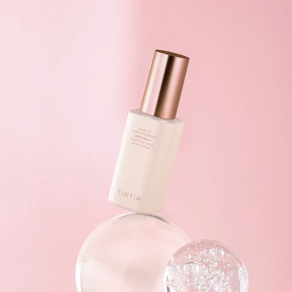 De afbeelding toont een fles "TIRTIR MASK FIT TONE UP ESSENCE" met SPF30 PA++ tegen een roze achtergrond, wat een gevoel van zachtheid en vrouwelijkheid geeft.