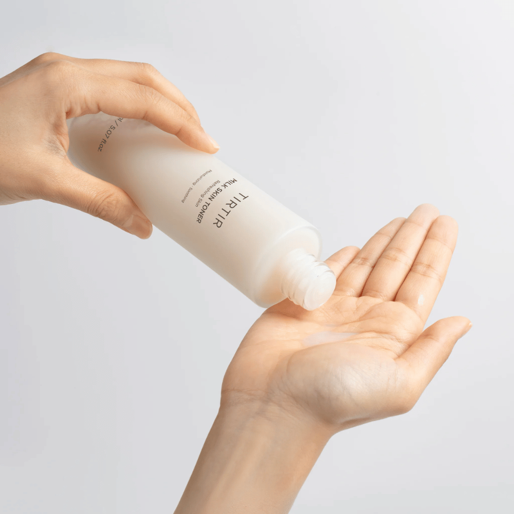 De afbeelding toont een persoon die een melkachtige vloeistof uit een fles met het label "TIRTIR Milk Skin" in de palm van de hand giet. Het product lijkt een huidtoner of vochtinbrengende lotion te zijn.