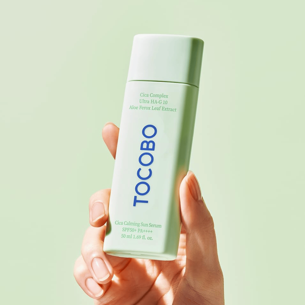 Hand houdt TOCOBO Cica Calming Sun Serum met mintgroene verpakking en blauwe tekst tegen lichtgroene achtergrond.