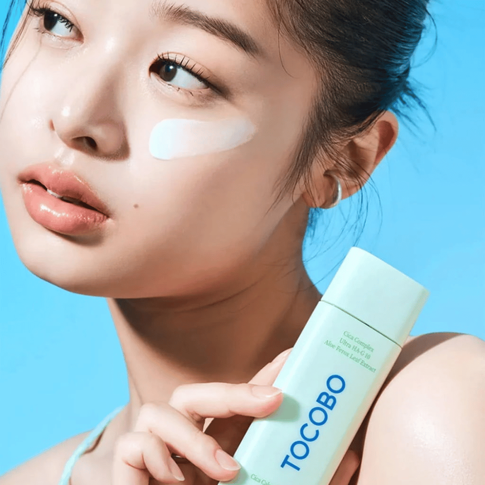 Model met zonnebrandcrème onder het oog houdt TOCOBO Cica Calming Sun Serum in mintgroene verpakking tegen blauwe achtergrond.