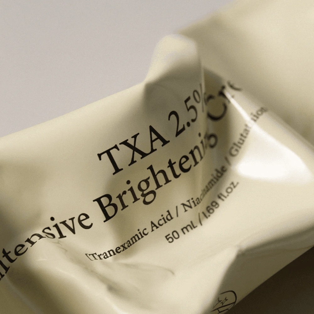 Close-up van AXIS - Y TXA 2.5% Intensive Brightening Cream waarbij het zachte en minimalistische tubedesign centraal staat. De afbeelding benadrukt de productnaam, Tranexamic Acid en Niacinamide, evenals de elegante beige verpakking.