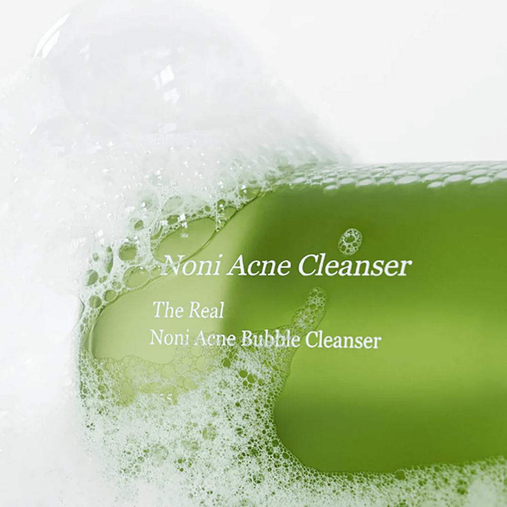 De fles met The Real Noni Acne Bubble Cleanser staat centraal en is omgeven door overvloedig schuim. De frisse groene kleur duidt op natuurlijke ingrediënten, perfect voor het reinigen en bestrijden van acne. Een ideaal product voor een schone en heldere huid.