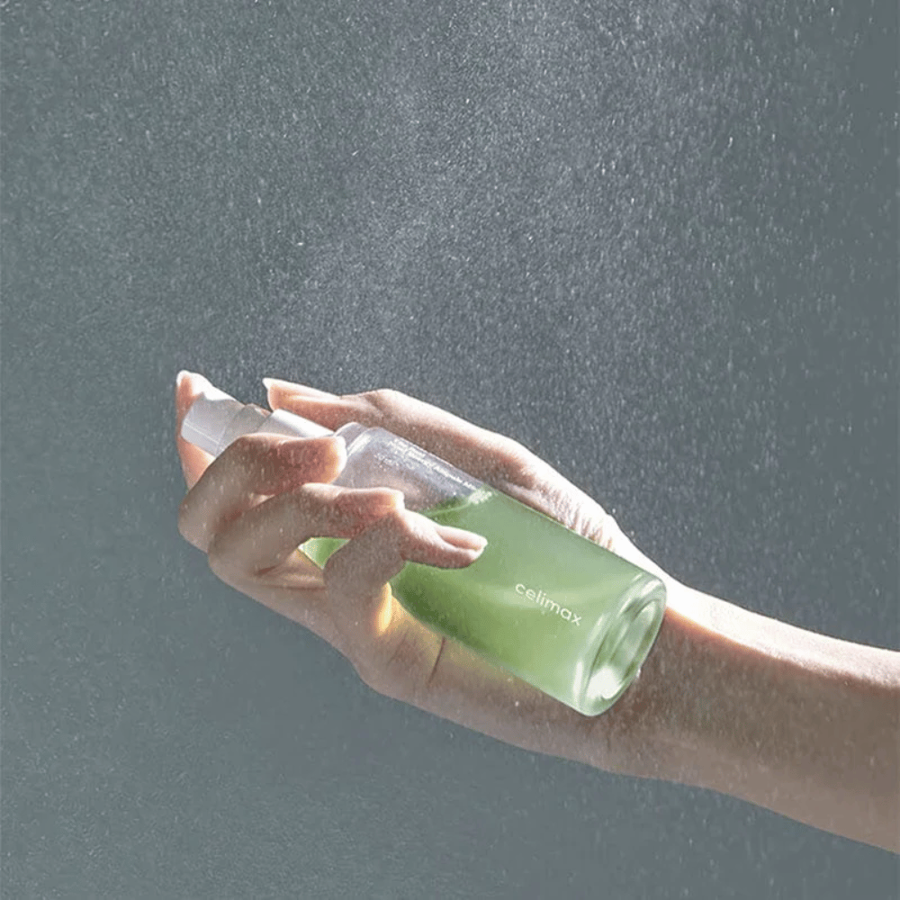 Een hand houdt Celimax Noni Mist spuitfles vast en brengt het product aan in een fijne, gelijkmatige spray op de huid. De fles is groen en het product wordt gepromoot als "The Real Noni Energy Ampule Mist" om energie en hydratatie aan de huid te geven.