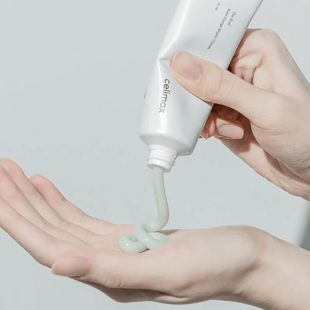 Een hand houdt een witte tube met Celimax Noni Energy Repair Cream, en de crème wordt in een gelijkmatige straal op de handpalm geperst. De crème is lichtgroen en wordt gepromoot om de huid te voeden en te herstellen.
