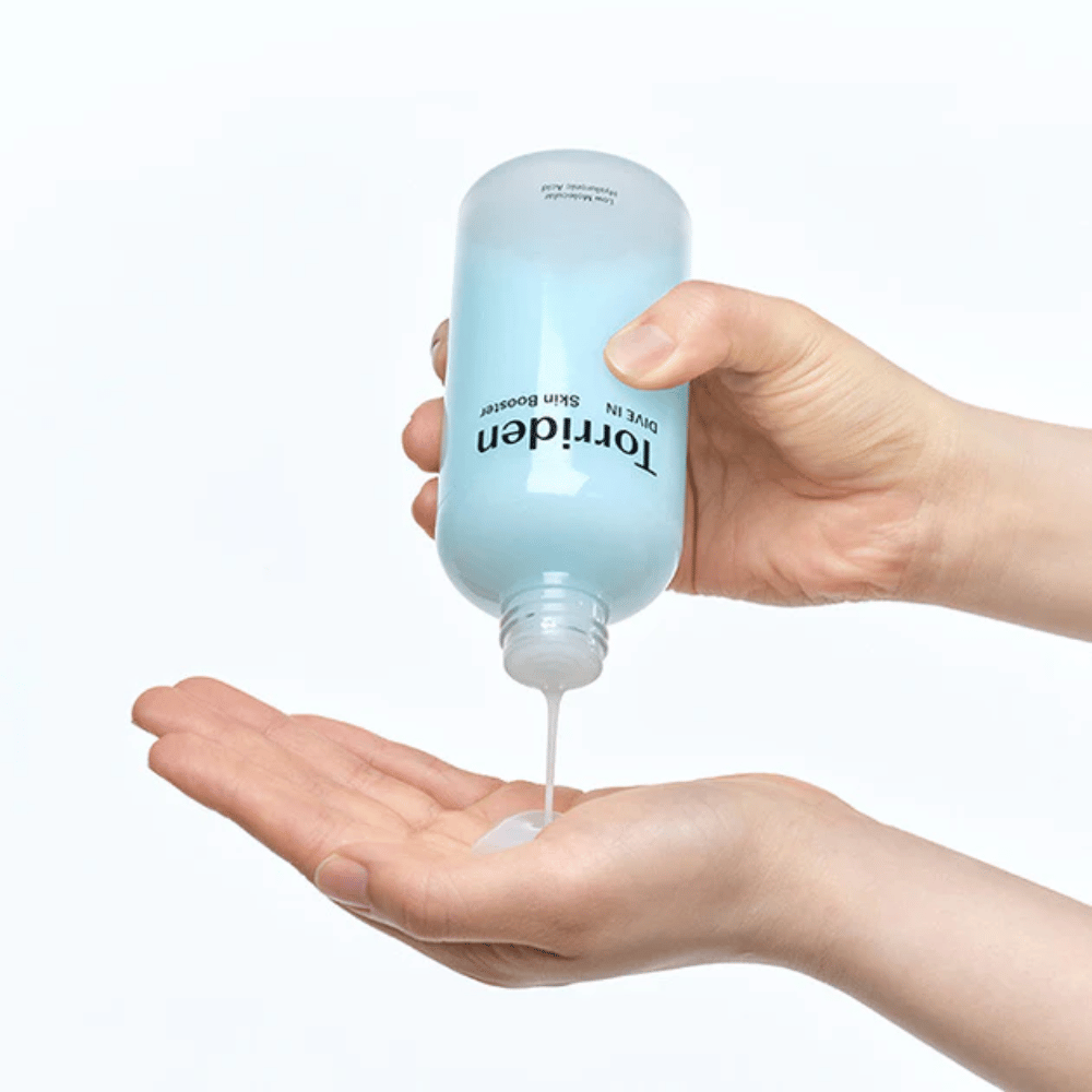 Handen die een fles met huidverzorgingsproduct vasthouden en een heldere vloeistof in de handpalm doseren. De fles is geëtiketteerd met "Torriden - DIVE-IN Low Molecule Hyaluronic Acid Skin Booster".
