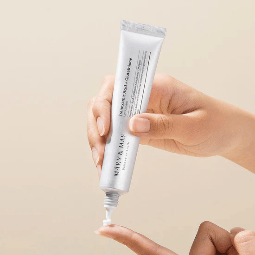 Een hand houdt een tube MARY & MAY "Tranexamic Acid + Glutathione Eye Cream". De tube is wit en het ontwerp is schoon en eenvoudig, wat de kalmerende en verhelderende eigenschappen van het product voor het ooggebied benadrukt. Het product wordt voorzichtig aangebracht, met focus op precisie en efficiëntie.