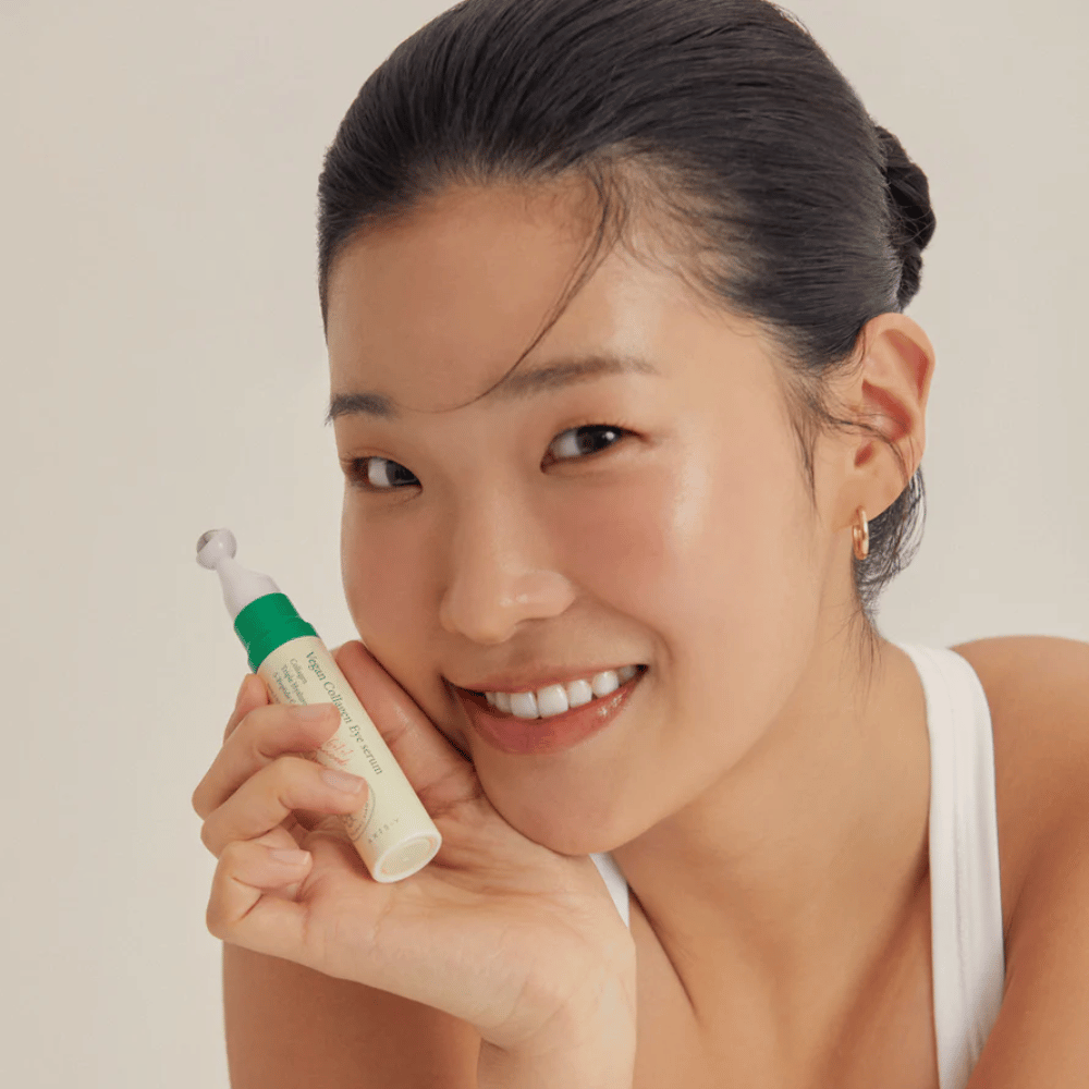 Een vrouw met opgestoken haar en gouden oorbellen houdt een AXIS-Y Vegan Collagen Eye Serum 10ml dicht bij haar gezicht terwijl ze zachtjes glimlacht. Het serum is ontworpen om te hydrateren en rimpels rond de ogen te verminderen, speciaal ontwikkeld voor de gevoelige huid.