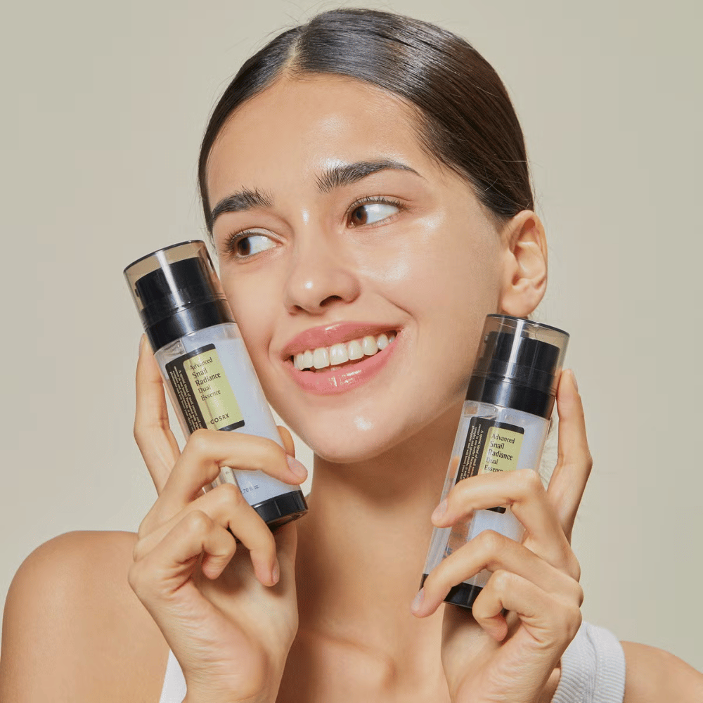 Lachende vrouw met glanzende huid houdt twee flessen COSRX Advanced Snail Radiance Dual Essence 80 ml dicht bij het gezicht tegen een lichte achtergrond.