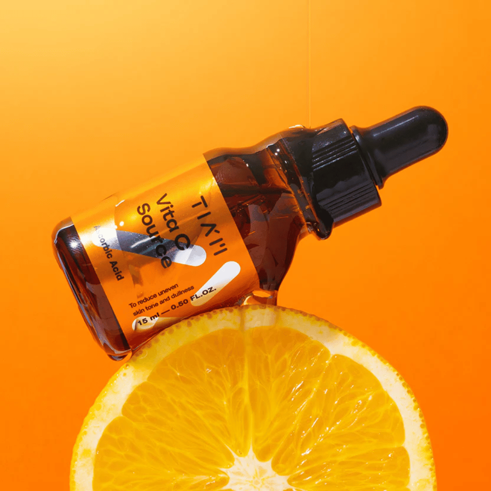 TIA'M Vita C Source-fles geplaatst tegen een sinaasappel en een oranje achtergrond die de frisheid en antioxidatieve werking van vitamine C symboliseert.