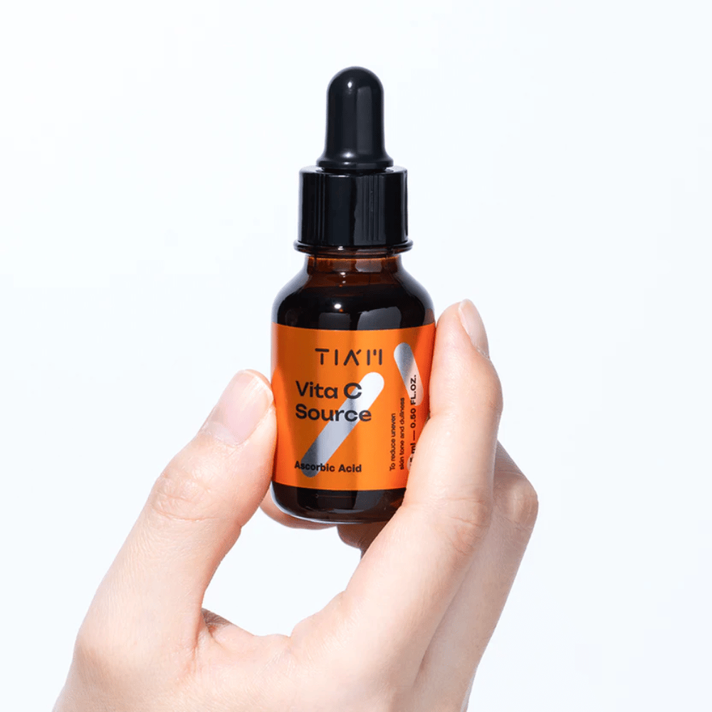Hand houdt TIA'M Vita C Source, een vitamine C-serum in een bruine glazen fles met pipet. Het product is ontworpen om de huidtint te egaliseren en glans te geven.